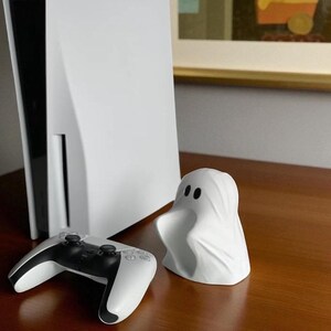May include: A white PlayStation 5 console and controller are displayed next to a small, white ghost figurine. The controller is black and white. The ghost figurine has two black eyes. The items are on a brown surface.