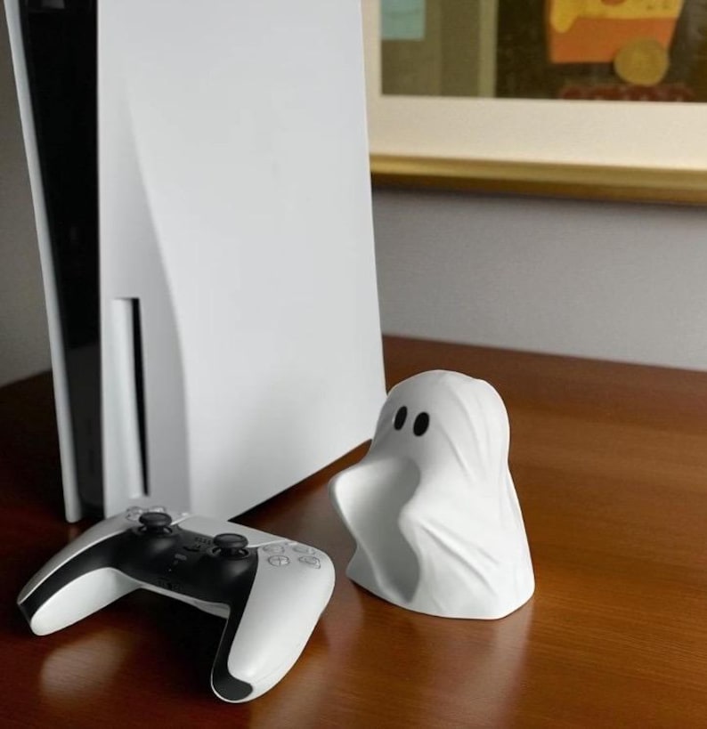 May include: A white PlayStation 5 console and controller are displayed next to a small, white ghost figurine. The controller is black and white. The ghost figurine has two black eyes. The items are on a brown surface.