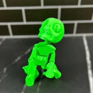 May include: A bright green 3D printed skeleton figurine with a comical expression. The skeleton is posed in a sitting position with its arms and legs bent.