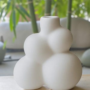May include: A white ceramic vase with a distinctive, rounded bubble design. The vase has a narrow neck and a series of spherical shapes that form its body. The background features green foliage, suggesting an indoor or outdoor setting.