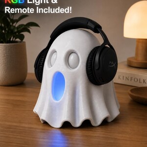 May include: A white ghost-shaped speaker with a blue light emanating from its mouth. Black headphones rest on the speaker's head. The text "RGB Light & Remote Included!" is visible. The speaker is on a wooden surface.