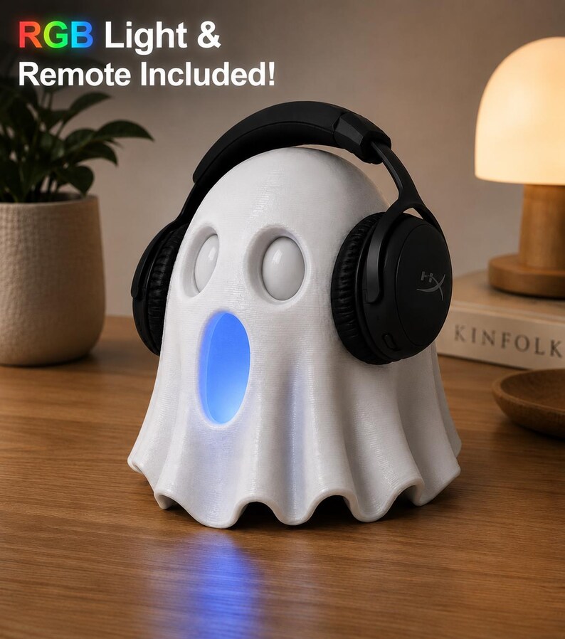 May include: A white ghost-shaped speaker with a blue light emanating from its mouth. Black headphones rest on the speaker's head. The text "RGB Light & Remote Included!" is visible. The speaker is on a wooden surface.