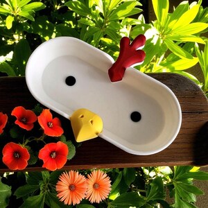 Op de afbeelding: Een witte keramische plantenbak met twee zwarte drainagegaten, een rood decoratief stuk en een gele snavelvormige accent. De plantenbak staat tegen een achtergrond van groen gebladerte en fel oranje bloemen.