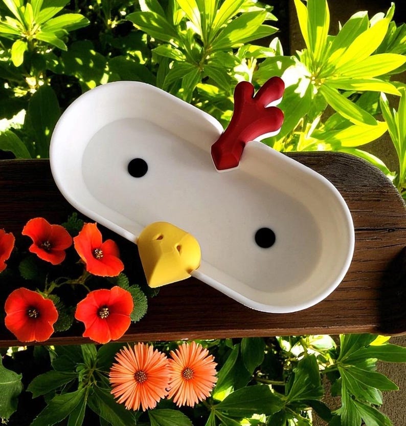 Op de afbeelding: Een witte keramische plantenbak met twee zwarte drainagegaten, een rood decoratief stuk en een gele snavelvormige accent. De plantenbak staat tegen een achtergrond van groen gebladerte en fel oranje bloemen.