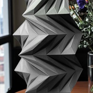 May include: A geometric, gray vase with a unique, angular design. The vase has a textured surface and is placed on a wooden table. Colorful flowers are in the background, adding a vibrant contrast to the vase's neutral tones. The vase is approximately 12 inches tall.