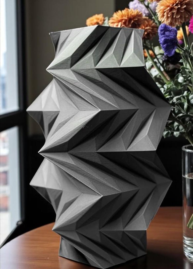 May include: A geometric, gray vase with a unique, angular design. The vase has a textured surface and is placed on a wooden table. Colorful flowers are in the background, adding a vibrant contrast to the vase's neutral tones. The vase is approximately 12 inches tall.