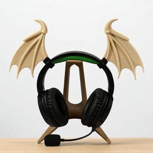 May include: Black gaming headphones with a green accent and attached microphone, on a tan headphone stand with bat wing accents. The stand is made of wood and has a three-legged design.