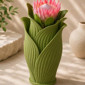 May include: A green, leaf-shaped vase holds a vibrant pink protea flower with green leaves. The vase has a textured surface and a round base. A neutral background highlights the flower and vase.
