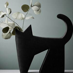 May include: A black cat-shaped vase with a textured surface. The vase has a tall, upright form with a pointed ear and a curved tail. The vase holds a sprig of eucalyptus leaves with round, silvery-green leaves. The background is a muted teal color.