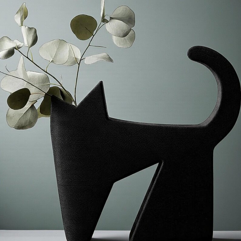 May include: A black cat-shaped vase with a textured surface. The vase has a tall, upright form with a pointed ear and a curved tail. The vase holds a sprig of eucalyptus leaves with round, silvery-green leaves. The background is a muted teal color.