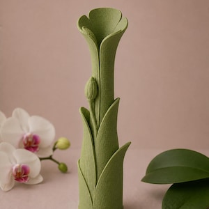 以下が含まれることがあります： 緑色の、花の形をした花瓶で、表面はざらざらしています。花瓶は細長い首と、咲いている花のように広がった上部を持っています。つぼみが側面にあります。花瓶は、ソフトピンクの背景に置かれています。