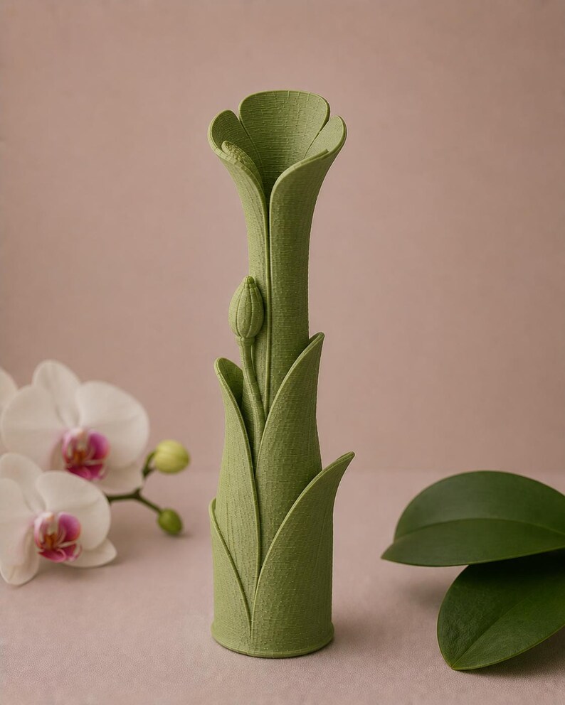 以下が含まれることがあります： 緑色の、花の形をした花瓶で、表面はざらざらしています。花瓶は細長い首と、咲いている花のように広がった上部を持っています。つぼみが側面にあります。花瓶は、ソフトピンクの背景に置かれています。