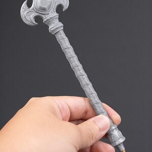 May include: A gray pen with an ornate, decorative design. The pen features a detailed, textured handle and a stylized top with a central gem-like element. The pen is held in a hand against a dark background.