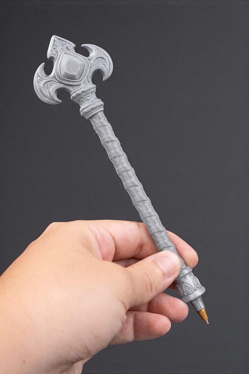 May include: A gray pen with an ornate, decorative design. The pen features a detailed, textured handle and a stylized top with a central gem-like element. The pen is held in a hand against a dark background.