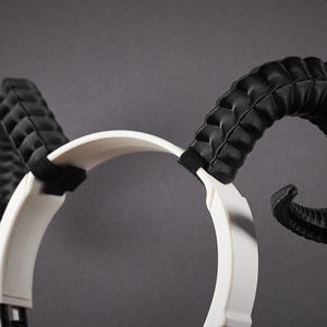May include: A white headband with black ram horns. The horns are curved and textured, with a quilted pattern. The headband is made of a smooth material, and the horns are attached with black straps.