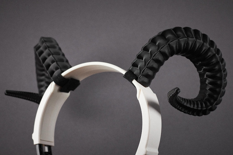 May include: A white headband with black ram horns. The horns are curved and textured, with a quilted pattern. The headband is made of a smooth material, and the horns are attached with black straps.