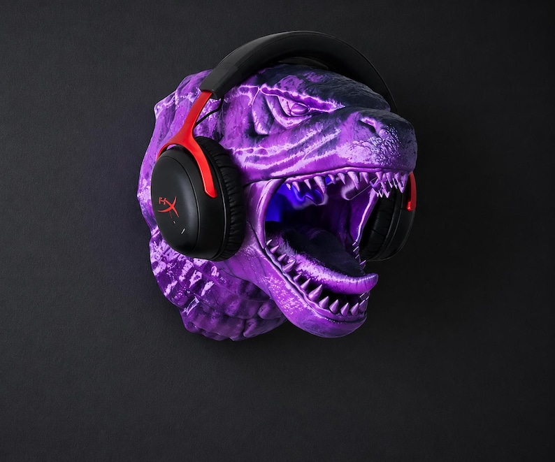 Może przedstawiać: Fioletowa rzeźba głowy Godzilli z czarno-czerwonym zestawem słuchawkowym do gier. Zestaw słuchawkowy ma logo HyperX. Rzeźba ma otwartą paszczę z widocznymi zębami i świecącym niebieskim wnętrzem. Tło jest czarne.