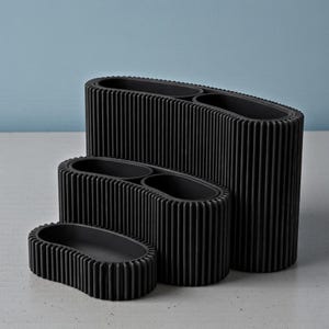 May include: Three black, ribbed, oval-shaped planters of varying sizes. The largest planter has two interior compartments. The planters are arranged on a light gray surface against a light blue background.
