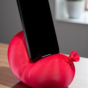 May include: A red phone stand shaped like a balloon, with a black smartphone resting in the center. The stand is made of a solid material and has a smooth texture. The phone is positioned upright.