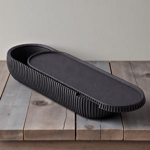 May include: A black, oval serving dish with a matching lid. The dish has a ribbed exterior and sits on a wooden surface. The lid is flat and fits inside the dish. This serving dish is suitable for serving food.