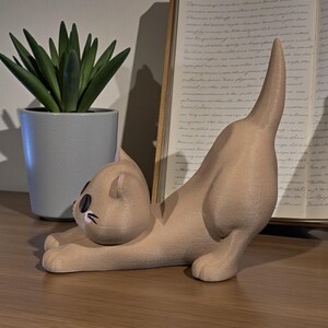 May include: A tan cat figurine in a stretching pose, with black eyes and a raised tail. The cat is on a wooden surface, with a potted succulent and an open book in the background.