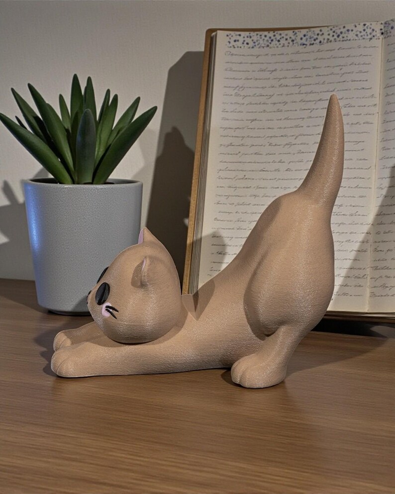 May include: A tan cat figurine in a stretching pose, with black eyes and a raised tail. The cat is on a wooden surface, with a potted succulent and an open book in the background.
