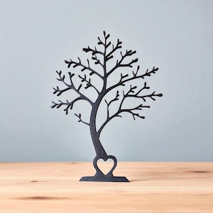 May include: Black metal tree-shaped jewelry holder with a heart cutout at the base. The tree has many branches and is displayed on a wooden surface against a light blue background. A decorative item for displaying necklaces, earrings, and bracelets.