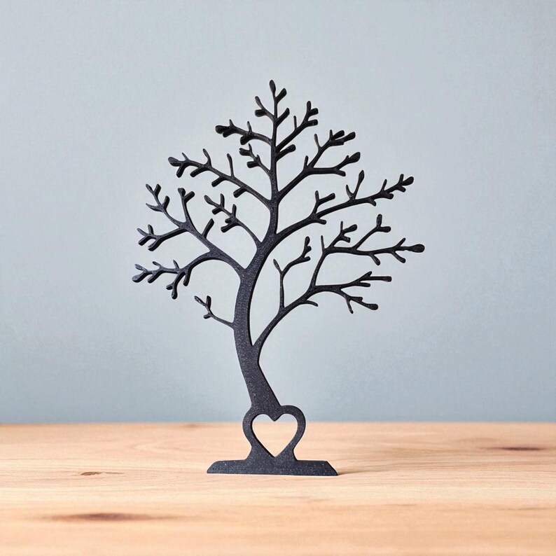 May include: Black metal tree-shaped jewelry holder with a heart cutout at the base. The tree has many branches and is displayed on a wooden surface against a light blue background. A decorative item for displaying necklaces, earrings, and bracelets.