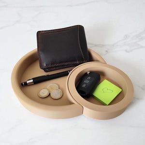 Puede incluir: Una bandeja beige de dos partes con una cartera de cuero marr&oacute;n, un bol&iacute;grafo negro, monedas, un llavero de coche y una nota adhesiva verde. La bandeja est&aacute; dise&ntilde;ada para objetos de uso diario.