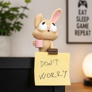 May include: A tan rabbit figurine with one upright ear and one folded ear, holding a small pink mug. The figurine features a brown clip on its back, currently holding a square yellow sticky note with the handwritten text "DON'T WORRY" in black ink.