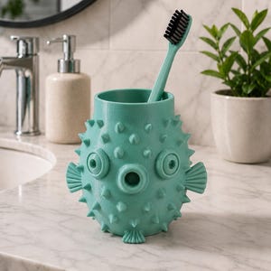 May include: A teal toothbrush holder shaped like a pufferfish, with a matching teal toothbrush. The holder has a textured surface with protruding spikes, eye-like features, and fin details. The toothbrush has black bristles.