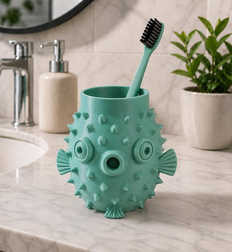 May include: A teal toothbrush holder shaped like a pufferfish, with a matching teal toothbrush. The holder has a textured surface with protruding spikes, eye-like features, and fin details. The toothbrush has black bristles.