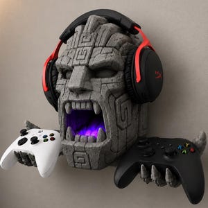 May include: A stone-textured wall mount shaped like a roaring face with geometric carvings, wearing black and red over-ear headphones. The figure has two stone hands extending outward, each holding a wireless game controller, one white and one black.