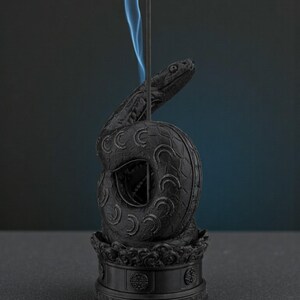 May include: Black incense holder in the shape of a coiled snake with a detailed design. The snake's head is raised, and blue smoke rises from the incense stick. The holder sits on a decorative base.
