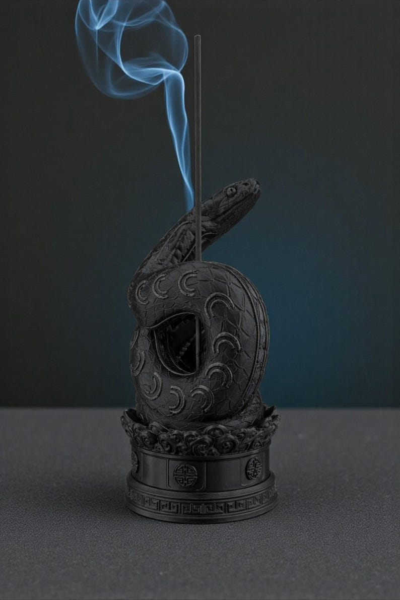 May include: Black incense holder in the shape of a coiled snake with a detailed design. The snake's head is raised, and blue smoke rises from the incense stick. The holder sits on a decorative base.