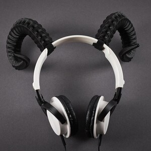 May include: White and black headphones with black, curved horn-shaped attachments on a grey background. The headphones have a white headband and black earcups. The horn attachments are black and textured.