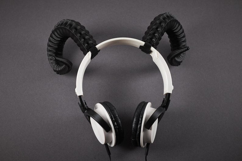 May include: White and black headphones with black, curved horn-shaped attachments on a grey background. The headphones have a white headband and black earcups. The horn attachments are black and textured.