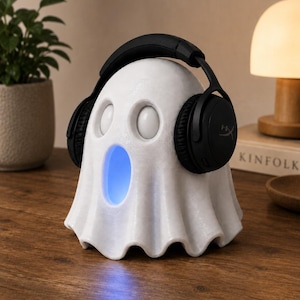May include: A white ghost-shaped speaker with a blue glowing mouth and black headphones. The speaker has two eye-shaped holes and a draped design. The headphones are black with the HyperX logo on the earcups.