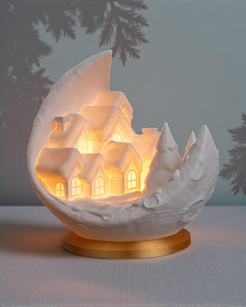 May include: A white ceramic Christmas village scene inside a crescent moon shape. The village features illuminated houses and trees, set on a gold-colored base. The background is a soft blue-green with a silhouette of a pine tree.