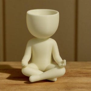 May include: A cream-colored figurine of a person in a seated yoga pose. The head is a large, open bowl shape. The arms are resting on the knees, and the legs are crossed. The figurine is on a wooden surface.