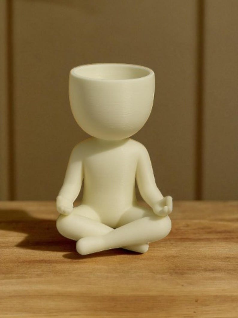 May include: A cream-colored figurine of a person in a seated yoga pose. The head is a large, open bowl shape. The arms are resting on the knees, and the legs are crossed. The figurine is on a wooden surface.