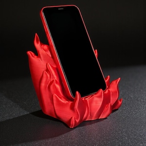 Puede incluir: Un tel&eacute;fono inteligente rojo descansa en un soporte para tel&eacute;fono rojo a juego. El soporte tiene un dise&ntilde;o en forma de llama, con formas curvas y fluidas. La pantalla del tel&eacute;fono es negra, contrastando con el rojo vibrante del tel&eacute;fono y el soporte.