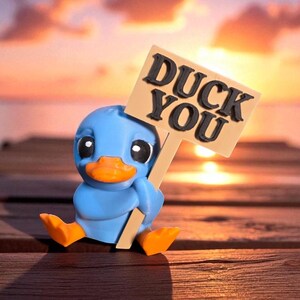 May include: A blue duck figurine with an orange beak and feet, holding a sign that reads "DUCK YOU". The sign is beige with black lettering. The duck is sitting on a wooden dock with a sunset in the background.
