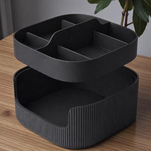 May include: A two-tiered, dark gray organizer. The top tray has multiple compartments, while the bottom section has a ribbed exterior. The organizer is designed to hold various items and sits on a wooden surface.