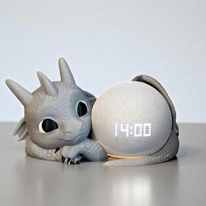 Puede incluir: Un soporte de altavoz gris con forma de drag&oacute;n que sostiene un altavoz esf&eacute;rico. El altavoz muestra la hora, 14:00, en d&iacute;gitos blancos. El drag&oacute;n tiene grandes ojos negros y una expresi&oacute;n amigable.