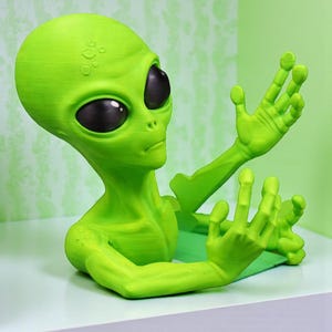 May include: A green alien figurine with large black eyes and outstretched arms. The alien is a vibrant green color and appears to be 3D-printed. The figurine is resting on a white surface, with a green background.