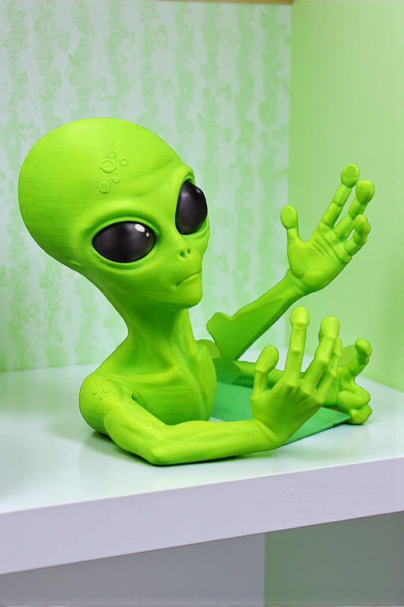 May include: A green alien figurine with large black eyes and outstretched arms. The alien is a vibrant green color and appears to be 3D-printed. The figurine is resting on a white surface, with a green background.