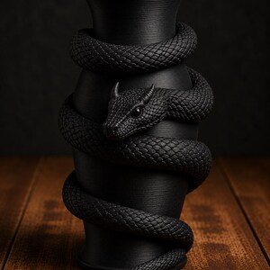 May include: A black vase with a textured surface, wrapped by a detailed, coiled snake. The snake's head is visible, with intricate scales and a menacing gaze. The vase sits on a wooden surface.