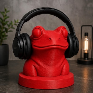 May include: A red frog-shaped headphone stand with black headphones. The stand is a vibrant red color and has a textured surface. The headphones are black and sit on the frog's head. The stand is on a round base.