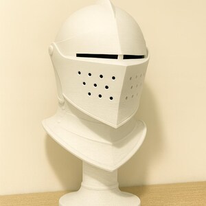 May include: A white knight's helmet sculpture with a visor featuring small circular holes. The helmet is mounted on a white pedestal, creating a bust-like appearance. The sculpture is made of a textured material.